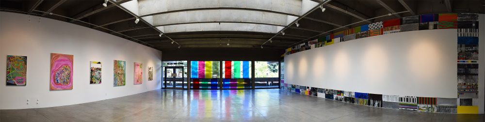 Panorama view of art gallery. On the left wall are six multicolored large abstract paintings; on the right wall are smaller multicolored abstract paintings that form a large rectangle. At center is a wall of glass windows that look out to greenery, with seven large bands of colored vinyl installed vertically across the glass.