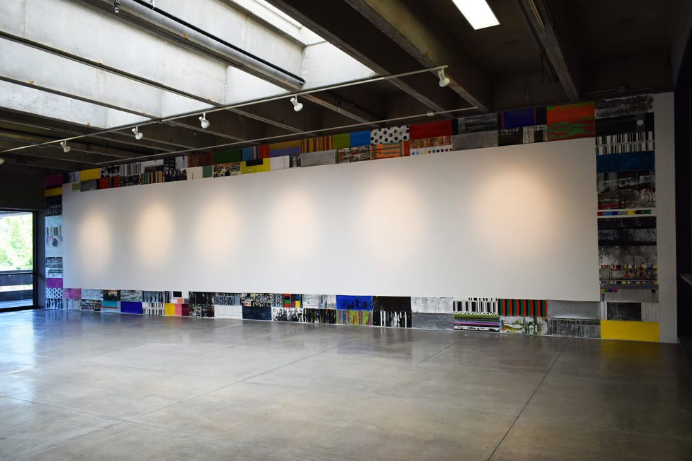 Gallery wall has an installation of 98 small paintings on paper, each measuring 11 by 30 inches. The paintings are directly adjacent to one another so as to form a rectangle at the perimeter of the wall; they form a large rectangle that touches the edges of the wall at the ceiling, the floor, and to the left and right. The center of the wall is empty except for the circular shapes created by the lighting for the artwork. The paintings are multicolored and abstract.
