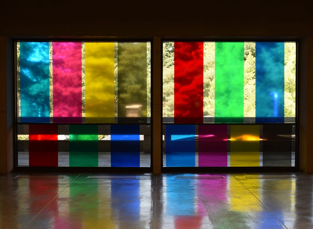 Artwork of vinyl rectangular colored decals on two glass walls. Colors include blue, red, green and yellow and are arranged as verticals. The trees and walkway are seen behind the glass.