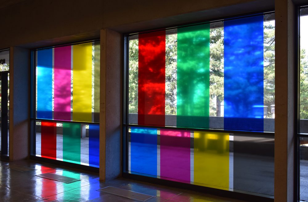 Artwork of vinyl rectangular colored decals on two glass walls. Colors include blue, red, green and yellow and are arranged as verticals. The trees and walkway are seen behind the glass.
