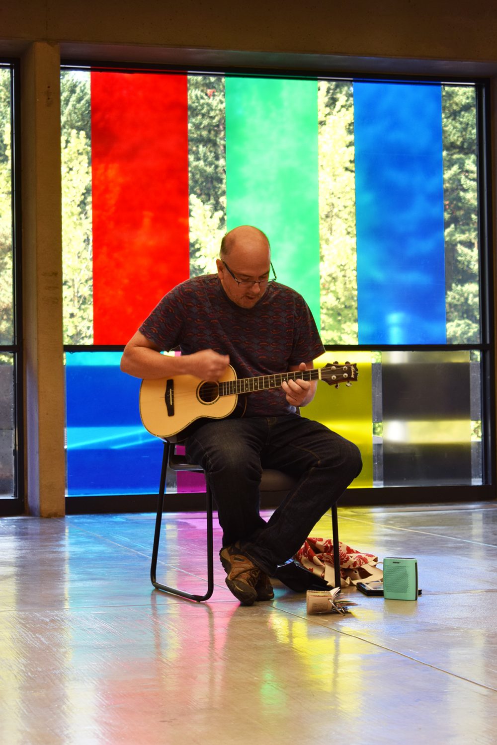 View of performance for the exhibition. A man is sitting on a chair playing a small guitar. On the floor beside him are objects. . Behind him is a wall of glass with large bands of blue, red, yellow and green vinyl.