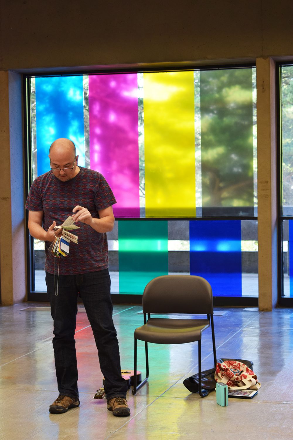 View of performance for the exhibition. A man stands to the left of an empty chair, a guitar and other objects are on the floor beside him. He is holding a large metal ring with painted cards on it and reading from the cards. Behind him is a wall of glass with large bands of blue, magenta, yellow and green vinyl.