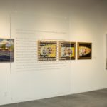 Installation view of gallery with a clear acrylic panel in the foreground and three artworks on the wall in the background. The acrylic panel has text printed on it. The artworks are paintings.
