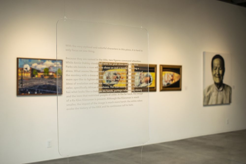 Installation view of gallery with a clear acrylic panel in the foreground and three artworks on the wall in the background. The acrylic panel has text printed on it. The artworks are paintings.
