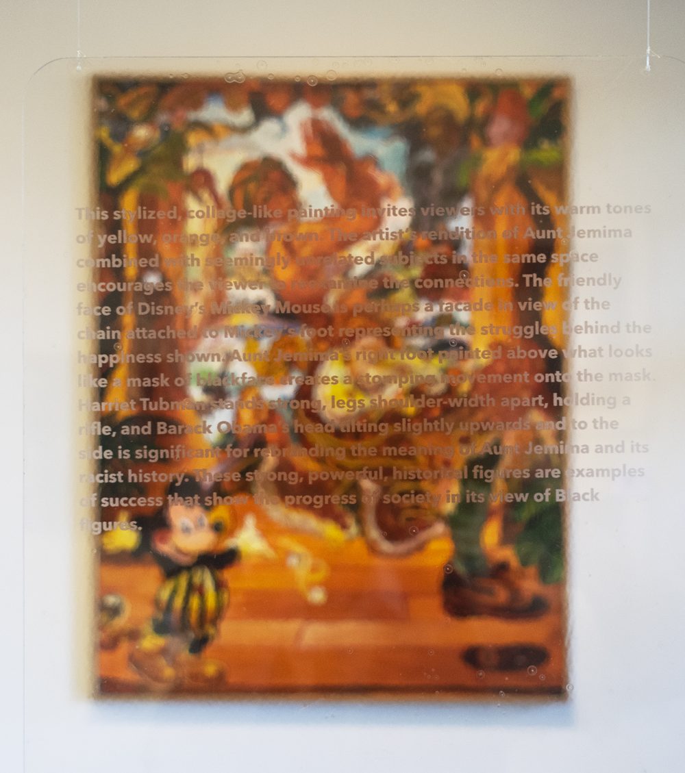 A painting of a woman dancing is hanging on a wall in the background. The painting is predominantly orange. In the foreground is text describing the painting, printed on a clear acrylic panel which is placed directly in front of it.