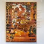 A painting of a woman dancing is hanging on a wall in the background. The painting is predominantly orange. In the foreground is text describing the painting, printed on a clear acrylic panel which is placed directly in front of it.