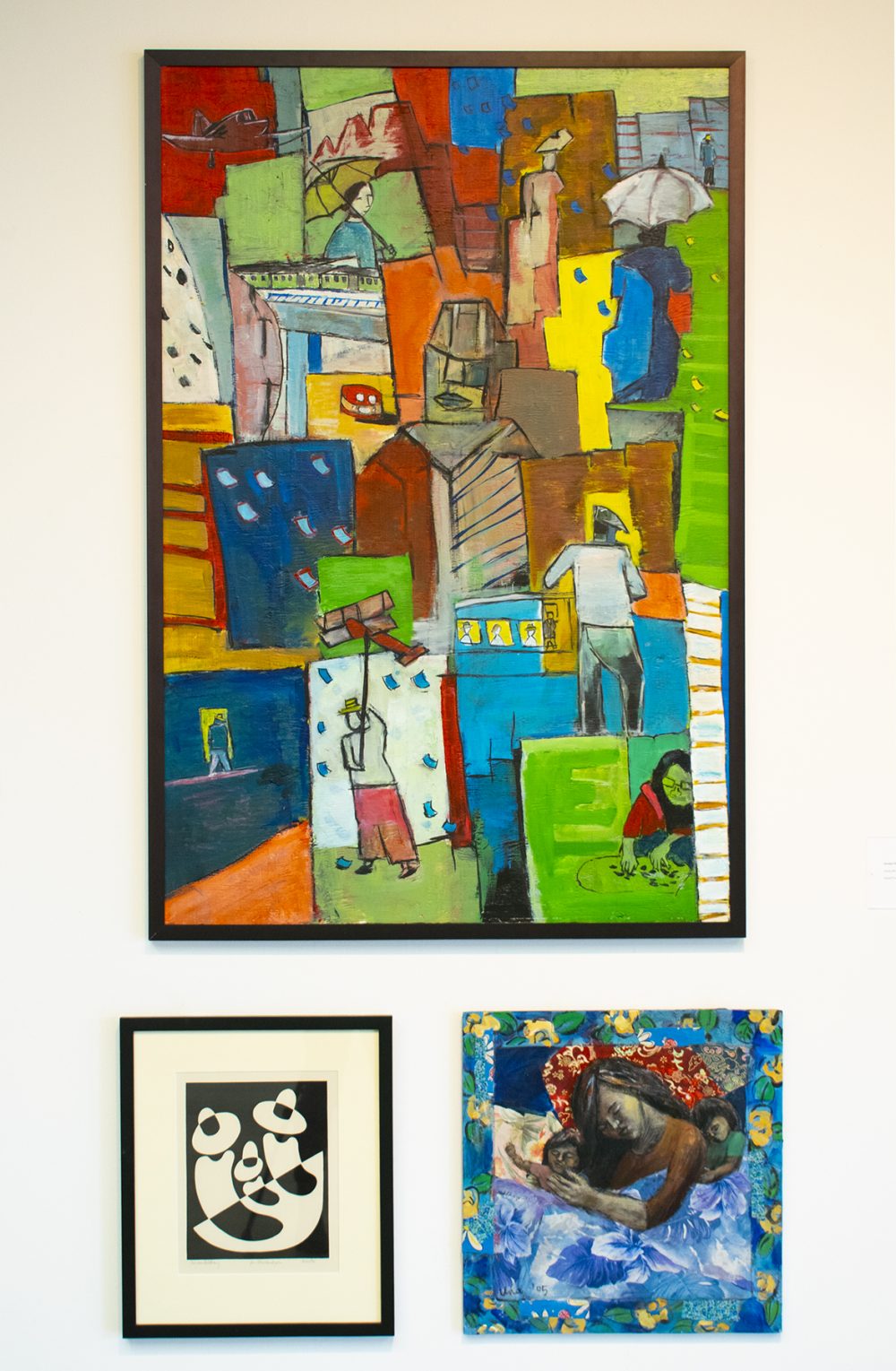 Three artworks hang on a wall. On top is a large multicolored, vertical, abstract painting depicting people in the street. On the bottom left is a small black and white print of three seated figures; on the bottom right a square painting of a woman sleeping in a bed with a child on either side of her. The painting is predominantly blue.