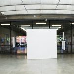 Panorama installation view of gallery with seven wall mounted artworks on the left and seven wall mounted artworks on the right. At center is a blank white wall and in the middle of the room are clear acrylic panels suspended from the ceiling. A video monitor is on a pedestal in the far right corner.