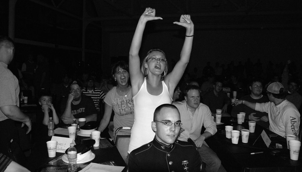Horizontal black and white photograph of a crowd of people sitting at tables with white paper cups and other objects on the tables. They are looking towards the right side of the photo. A young man in a forman marine uniform sits in the foreground; a young woman in a white shirt stands behind him with her hand raised above her head and cheers. Behind and to her left is another woman who stands and shouts. The background is dark.