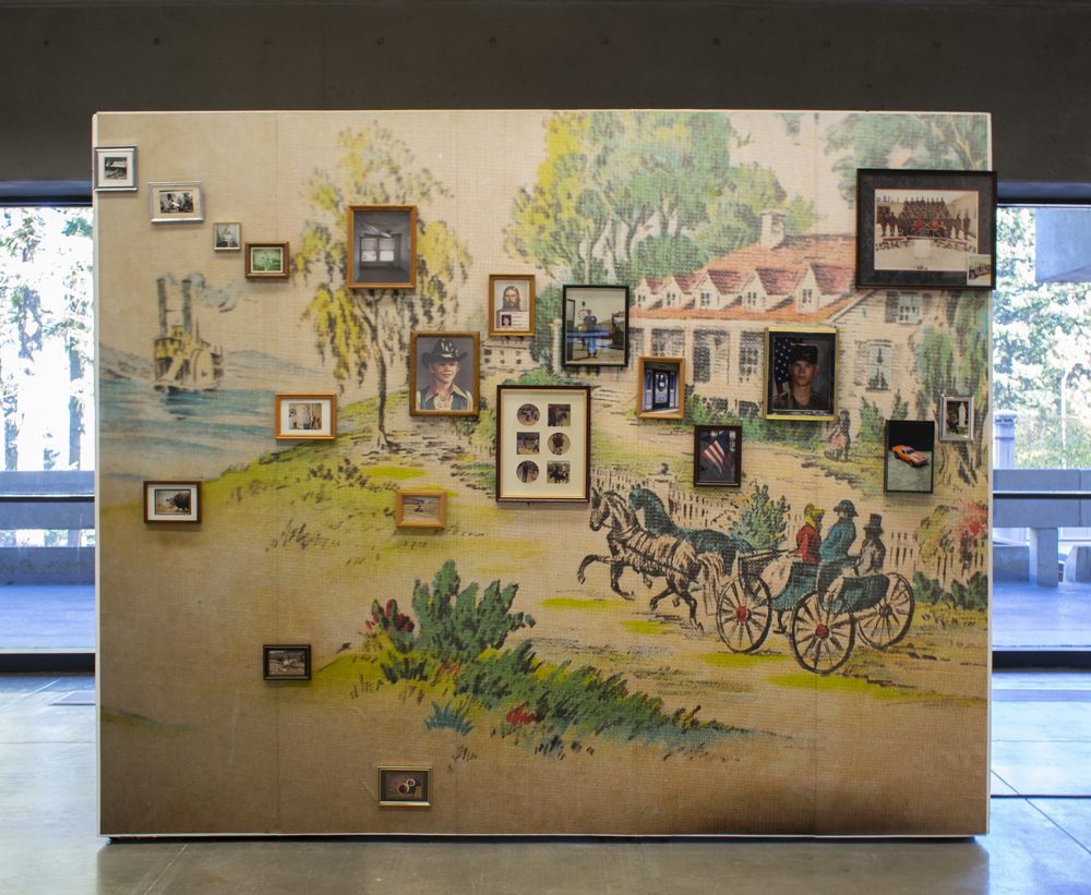 A freestanding wall in a gallery is in the foreground and floor to ceiling windows and the landscape outside are in the background. The wall has old fashioned, faded wallpaper covering it; the wallpaper depicts an antebellum era pastoral scene of three people in a horse-drawn carriage outside a large home. Installed on the papered wall are many small scale framed photos.