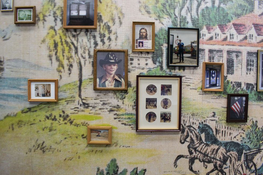 Several small scale framed photos are hung on a wallpapered wall. The photos faded and appear to be from a personal family album. The wallpaper is multicolored and depicts a pastoral scene with horses and a house on the right and grass and trees and a lake on the right.