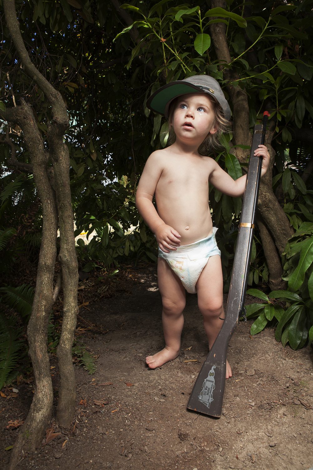 Vertical color photograph of a small boy, wearing only a diaper and a confederate army style hat, and holding a wooden replica of a civil war musket. He gazes up and to the left. He is standing on brown dirt and is surrounded by shrubs in the background. At the left of the photo are two slender tree trunks.