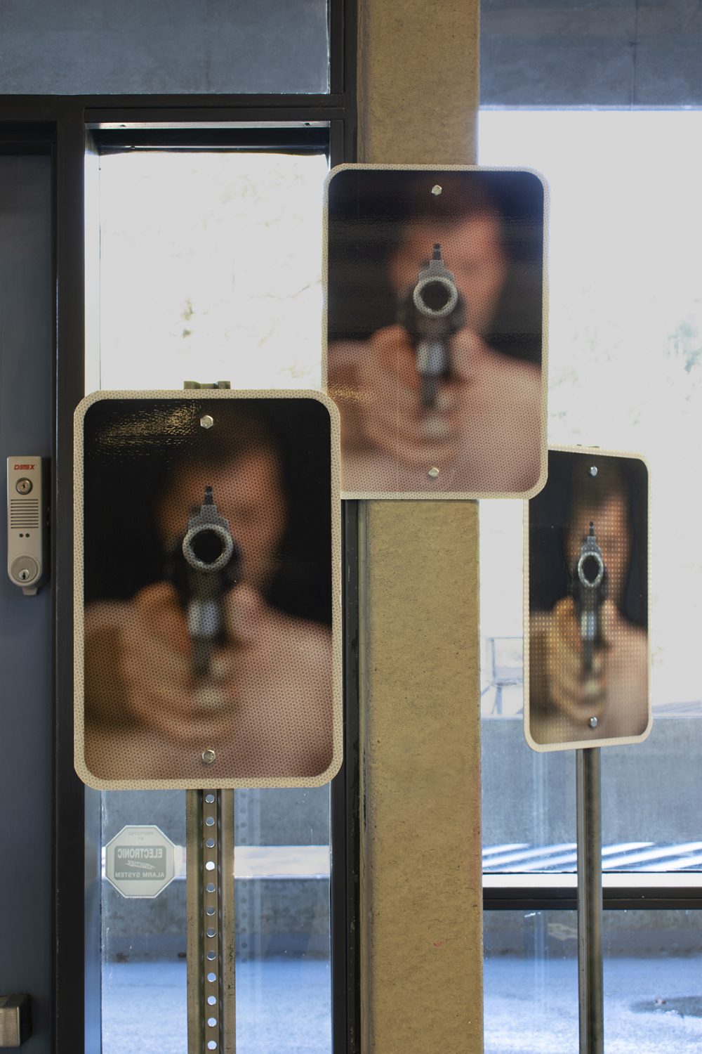 Three identical signs are in the foreground, each depicting the upper body of a man holding a large gun that is pointed at the viewer. In the background are floor to ceiling glass windows with a grey door with a lock on the right,and a concrete column at the center.