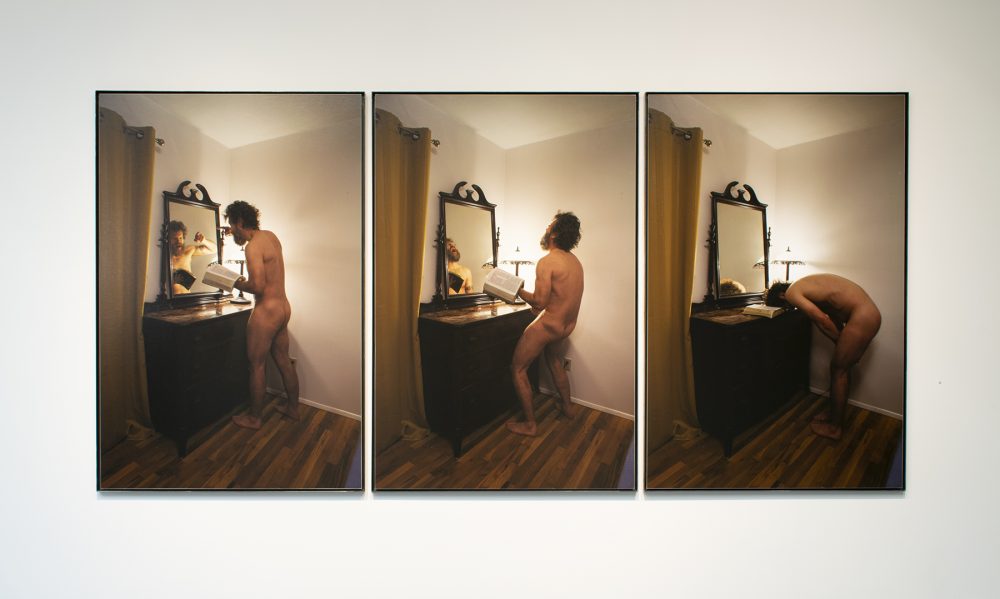Three large scale framed color photographs hang on a white wall. The images on each photo are sequential, and take place in the same setting. The photo on the left depicts a naked man, holding a bible, standing in front of a dresser with a mirror on it. He is pointing at himself in the mirror. The center photo shows the man with his head thrown back and his mirror image is yelling. The photo on the right shows the same man bent with his hands down and his head resting on the bible which is on the dresser.