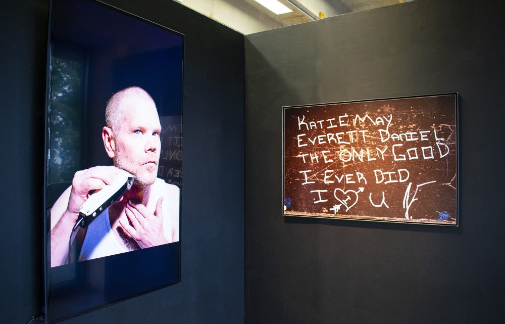 Installation view of a corner a gallery; the walls are black and meet at the center of the image. On the left is a video monitor with an image of a man with a clean shaven head who is shaving his chin with an electric razor. On the left wall is a photo of a black board with the words "Katie May Everett Daniel the only good I ever did" written on it in white block letters.