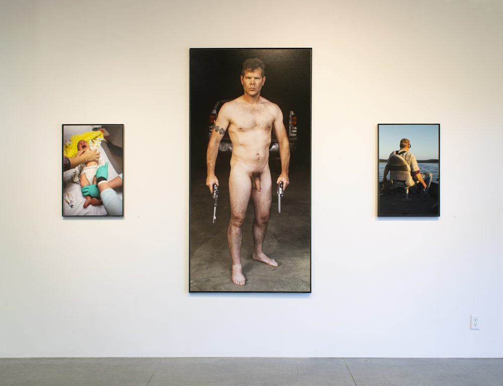 A gallery wall installation of three vertical color photographs, one large scale at the center and two smaller same sized ones to the left and right of it. The left photo shows a crying baby on a hospital table with two pairs of adult hands touching it. One pair of hands is wearing green hospital gloves. The center photo is of a naked man standing and looking at the viewer, each of his arms are held straight at his sides and he has a gun in each hand. The right photo is of a man sitting in a boat, with his back towards the viewer; in the background is a lake and sky