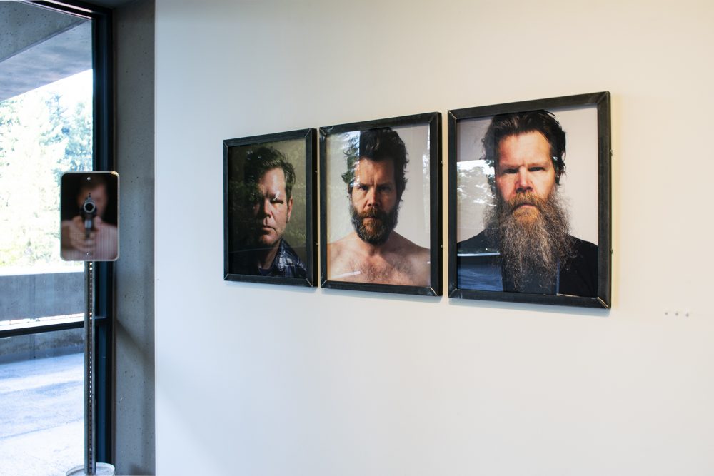 Installation view of the corner of a gallery, with a white wall on the left and a glass wall looking outside on the right. On the wall are three framed photographic portraits of men, each showing only the head and shoulders, and with the man looking straight at the viewer. In the corner, to the left is a sign on a metal post, the sign depicting a man pointing a large handgun at the viewer.