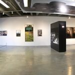 Panorama view of gallery showing an installation of large scale photographs. On the left is a freestanding wall with a photo of a pick up truck and a wall of smaller framed photos. On the right are two freestanding walls painted black and that have black and white photos of people on them. In the background is a white wall with several photos hanging on it. At the center of the back wall is a lifesize photo of a sign that says "Whiteville." Hanging horizontally on the ceiling at center right is panel with a photo of a car's underside on it.
