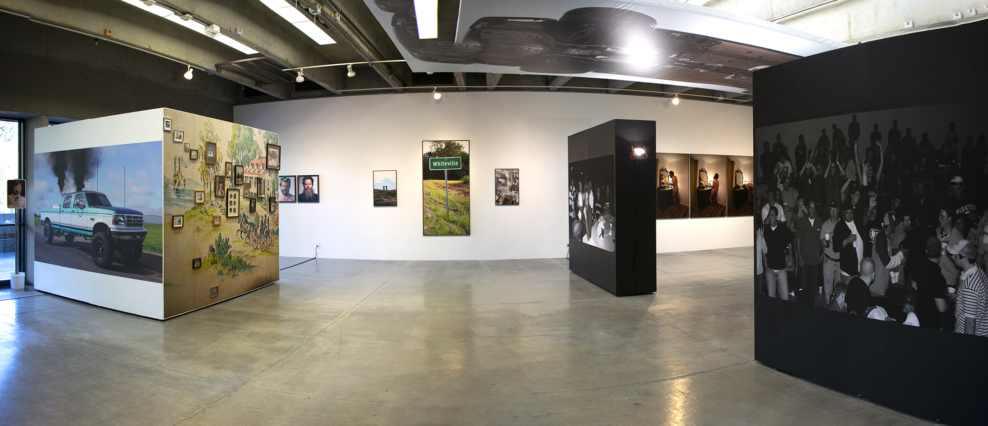 Panorama view of gallery showing an installation of large scale photographs. On the left is a freestanding wall with a photo of a pick up truck and a wall of smaller framed photos. On the right are two freestanding walls painted black and that have black and white photos of people on them. In the background is a white wall with several photos hanging on it. At the center of the back wall is a lifesize photo of a sign that says "Whiteville." Hanging horizontally on the ceiling at center right is panel with a photo of a car's underside on it.