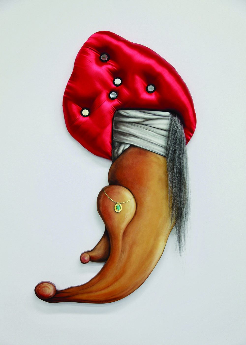 Mixed media sculpture hangs on a white gallery wall. The lower half of the artwork is painted wood and depicts a jeweled necklace and white wrapped fabric. The upper part of the piece is a padded turban like shape made of shiny red fabric, with white buttons imbedded in it. A tail like drape of artificial grey hair is draped on the right side of the object.