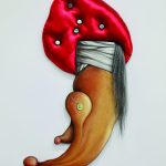 Mixed media sculpture hangs on a white gallery wall. The lower half of the artwork is painted wood and depicts a jeweled necklace and white wrapped fabric. The upper part of the piece is a padded turban like shape made of shiny red fabric, with white buttons imbedded in it. A tail like drape of artificial grey hair is draped on the right side of the object.