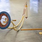 Large scale mixed media sculpture sits on a concrete floor in a gallery. The artwork is abstract and multicolored. On the right of the piece is a blue circle within which is an opening that is painted in trompe l'oeil style to resemble folds and flesh or bone like forms. The circle is attached to two pieces of bent metal, painted light yellow, which sit on the floor. At the other end of the bent metal is a pink metal wire form from which is suspended a white fabric baglike shape. On the bottom right of the sculpture is a long wooden pole that extends from the yellow metal curves across to a small orange stand.