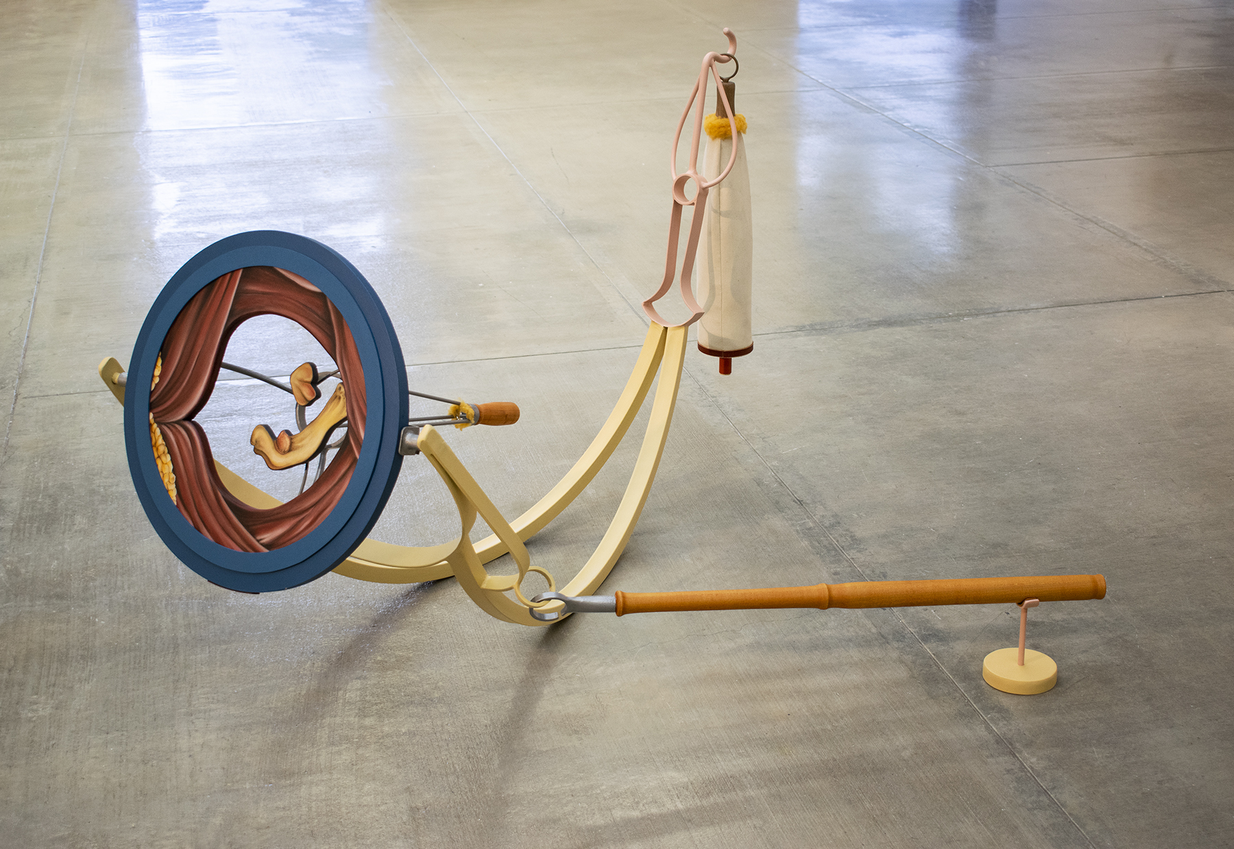 Large scale mixed media sculpture sits on a concrete floor in a gallery. The artwork is abstract and multicolored. On the right of the piece is a blue circle within which is an opening that is painted in trompe l'oeil style to resemble folds and flesh or bone like forms. The circle is attached to two pieces of bent metal, painted light yellow, which sit on the floor. At the other end of the bent metal is a pink metal wire form from which is suspended a white fabric baglike shape. On the bottom right of the sculpture is a long wooden pole that extends from the yellow metal curves across to a small orange stand.