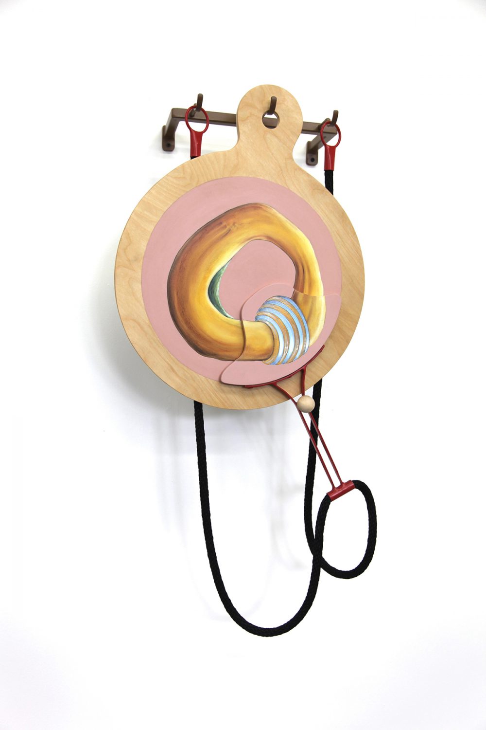 Large mixed media sculpture installed on a white gallery wall. A brown metal bar with three hooks is mounted to the wall on both ends, jutting out about 5 inches. A paddle shaped wood panel hangs from the center hook, and on it is a painting of a round yellow form on a pink circular background. A black rope with a red metal ring on each end hangs on the two other hooks.