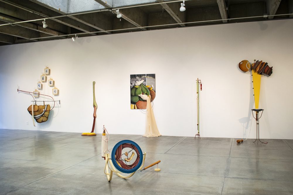 : Installation view of gallery shows five large scale mixed media sculptures hanging on a white wall in the background and one mixed media sculpture on the floor in the foreground. The artworks lean on the wall and/or sit on the floor and are abstract and multicolored.