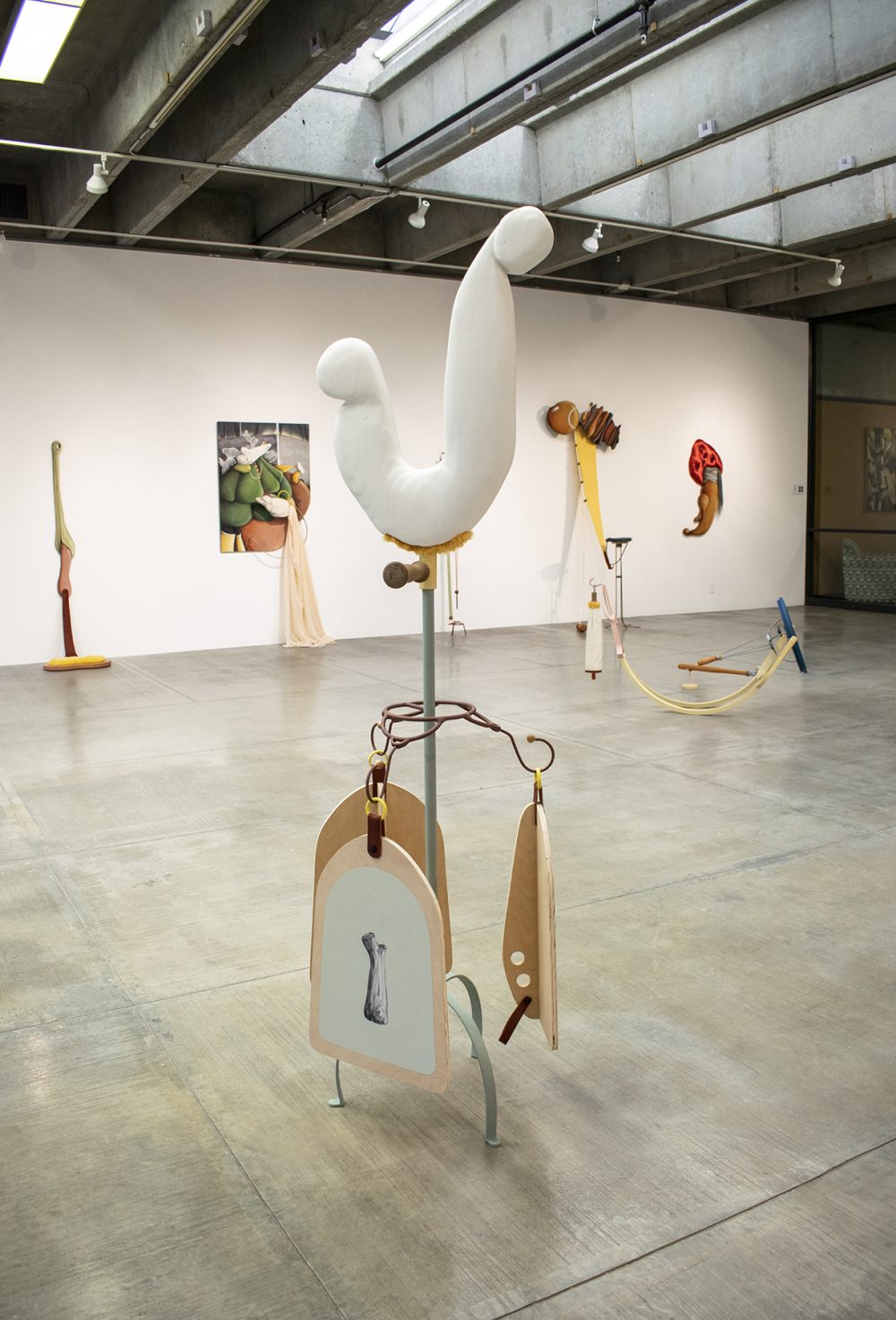 Installation view of exhibition shows large scale mixed media sculptures on the floor and wall of the gallery. Four wall mounted artworks are in the background, and on the right in the middle of the floor is an artwork that is close to the ground and made of bent metal, appearing as though it may rock. In the foreground is a tall sculpture with a central pipe from which hang three wooden paddle shapes with paintings on then. The piece stands on three metal legs, and at the top of the artwork is a white form in the shape of a "J" that is made of stuffed fabric. All the works are abstract and multicolored.