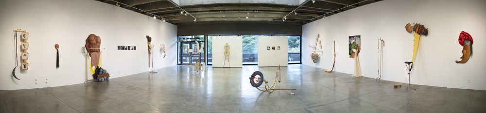 Panorama view of gallery. On the left and right walls are large scale mixed media sculptures that hang on the wall, some with elements that fall onto or stand on the floor. There are six artworks on the right wall and six artworks on the left wall. At the center in the background are two square walls in front of a glass wall that shows the landscape outside. The left back wall has a sculpture that stands on the floor and leans on the wall. The right back wall has three small paintings. On the floor in the middle of the gallery are two mixed media sculptures. All the artworks are multicolored and abstract. The materials used include, metal, wood, fabric, hair, leather, and paint, among other things.