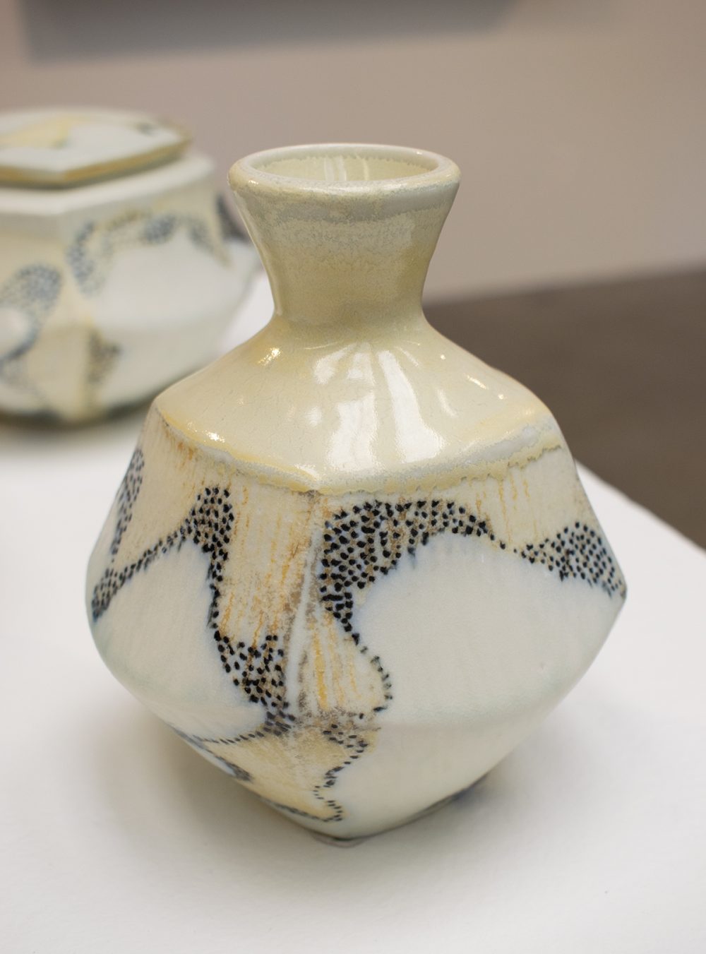 Ceramic vase like form sits on a white pedestal, with another ceramic object behind it on the upper left. The ceramic form is narrow at the bottom, widens in the middle, has narrow shoulders, becomes narrowest at its nect and widens again at the top where there is a round opening. The surface is glossy and the design is of bluish black dots that wander across the body of the vase with large areas of white abstract forms on the lower right and on the middle left. The rest of the vase is a yellowish beige with light streaks of orange in some areas.