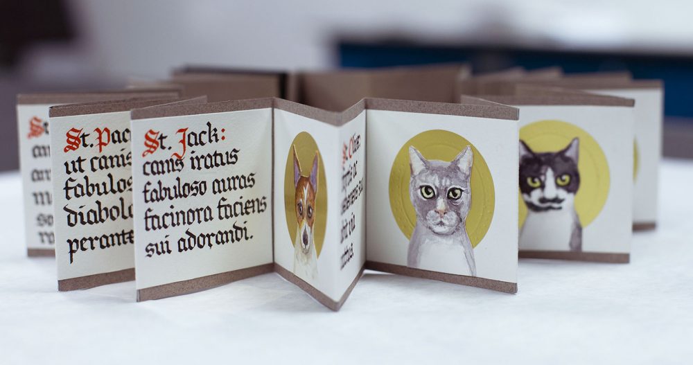 A small format accordion book is open and sitting on a white surface. The book is handpainted in watercolors, and has over a dozen pages, each 3 inches square. The pages on the left show calligraphic lettering in red and black. The pages on the right each portray an illustration of a cat's face in front of a gold circle. At the center of the image is the face of a dog in a gold circle. The book has is bordered on top and bottom with an eight of an inch strip of brown.