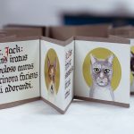 A small format accordion book is open and sitting on a white surface. The book is handpainted in watercolors, and has over a dozen pages, each 3 inches square. The pages on the left show calligraphic lettering in red and black. The pages on the right each portray an illustration of a cat's face in front of a gold circle. At the center of the image is the face of a dog in a gold circle. The book has is bordered on top and bottom with an eight of an inch strip of brown.