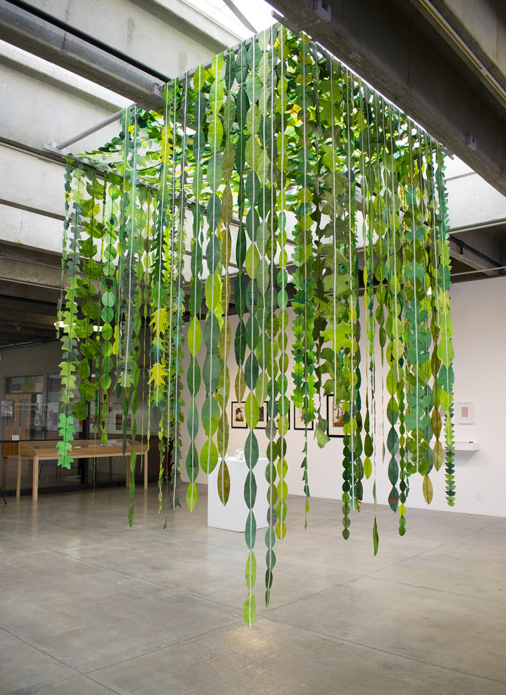 A sculptural installation hangs from the ceiling of a gallery, with a grey concrete floor below and other artworks on a white wall in the background. The installation consists of strips of white paper with green paper leaf shapes attached to each other end to end. These hang at different levels but do not reach the floor. Each strip is suspended from a wooden bar that is attached to the ceiling joists. The leaves create a small area for a viewer to stand under.