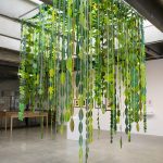 A sculptural installation hangs from the ceiling of a gallery, with a grey concrete floor below and other artworks on a white wall in the background. The installation consists of strips of white paper with green paper leaf shapes attached to each other end to end. These hang at different levels but do not reach the floor. Each strip is suspended from a wooden bar that is attached to the ceiling joists. The leaves create a small area for a viewer to stand under.