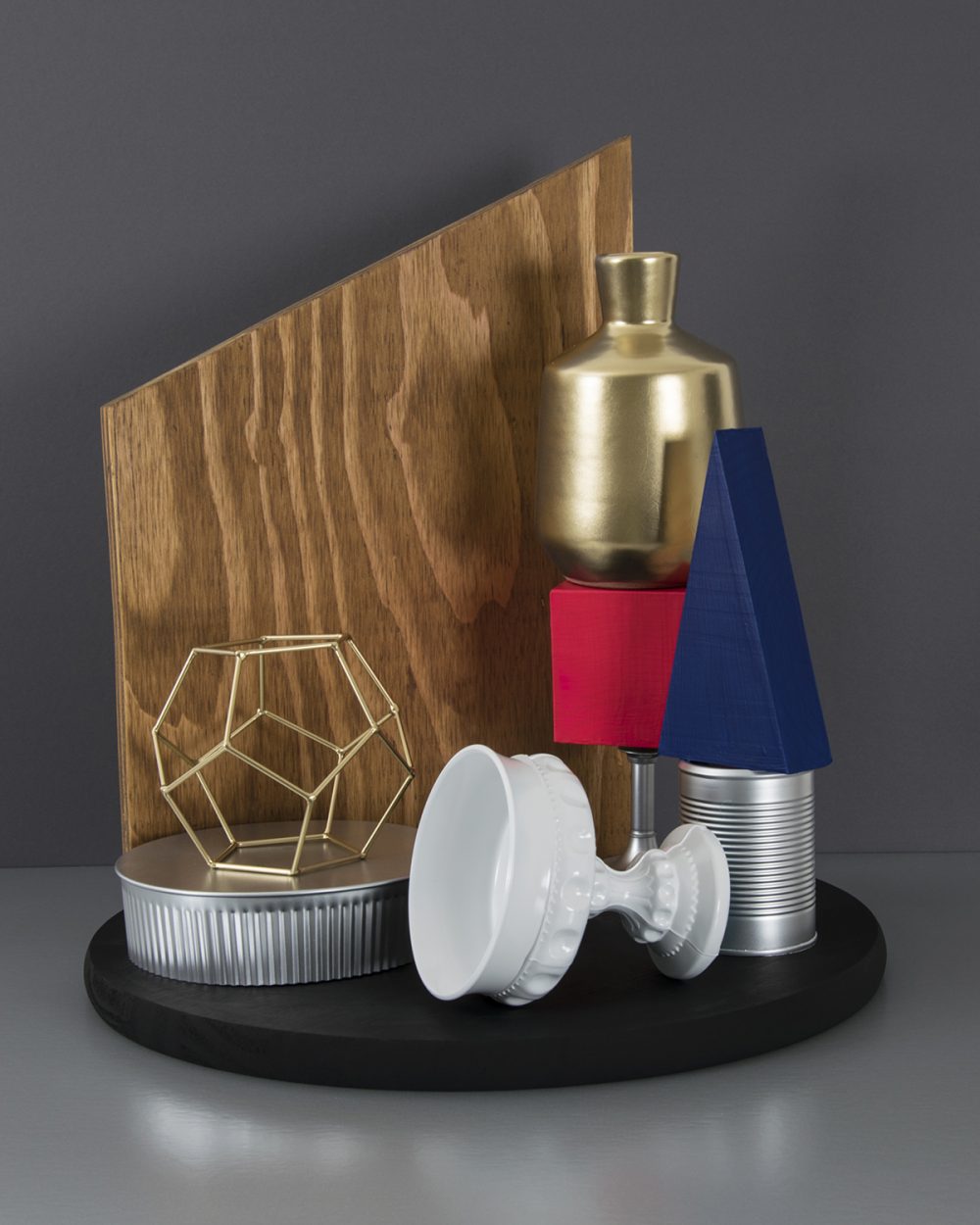 Digital photograph of a still life arrangement set on a gray reflective surface, with a darker gray background. A round black form serves as a base for a number of items: a silver tin can with an indigo triangle on top of it on the right; a silver stem with a red cube and gold vase beside that; a white goblet on its side in front of the tin can; on the left a ribbed silver form with a gold wire shape on top of it; behind on the center and left is a piece of wood plank, cut at an angle and sitting on end.