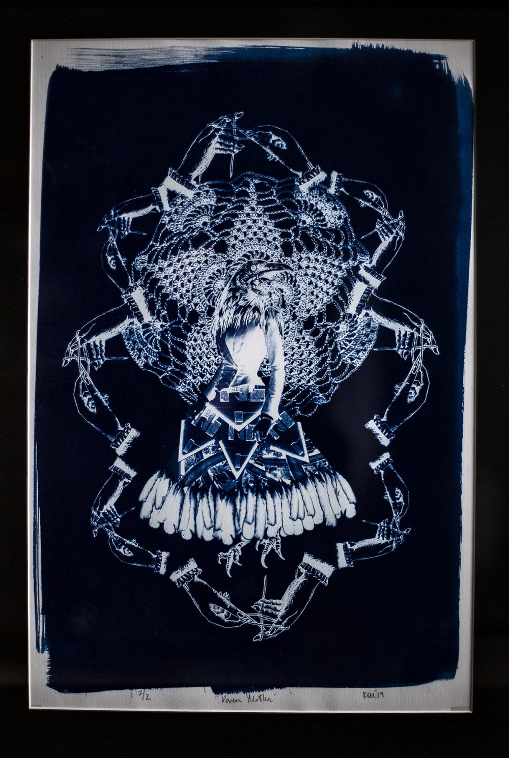 A cyanotype print in a black frame. A central female figure with a birds head, wears a skirt of feathers, and has bird claws for feet. A lace doilly is behind her, like a halo. Surrounding the iconic form is an undulating border or hands with knitting needles. The image is printed in tones of indigo and white, with a very dark blue in the background.