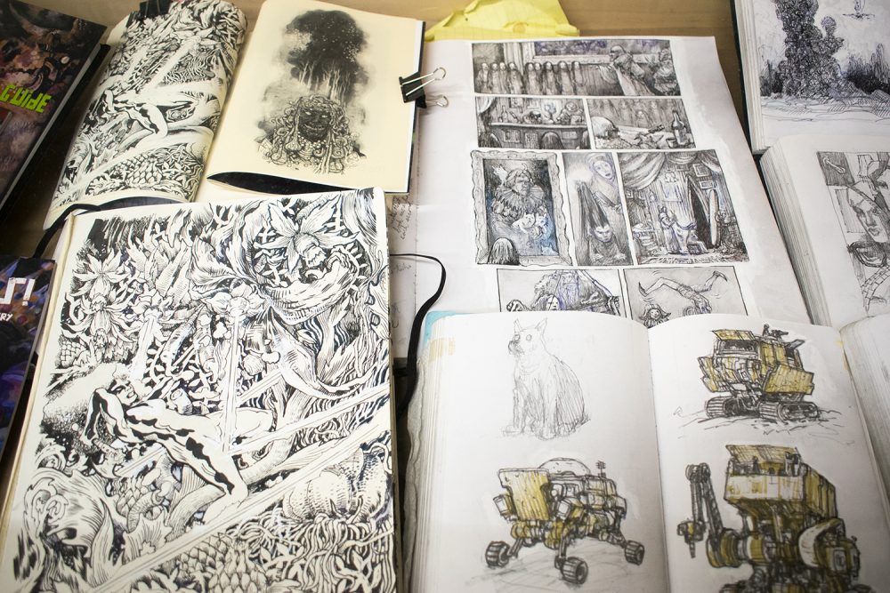 Open sketchbooks lay beside each other in a vitrine. On the lower right an open book shows two pages of ink drawings of an imaginary armored vehicle, and a pencil drawing of a dog. Above that is a sketchbook page with a gridded set of scenes layed out comic book style, depicting figures and interiors. On the lower left is a large ink drawing of a figure surrounded by fantastical foliage, and above that a smaller drawing of the head of a character with worm like hair.