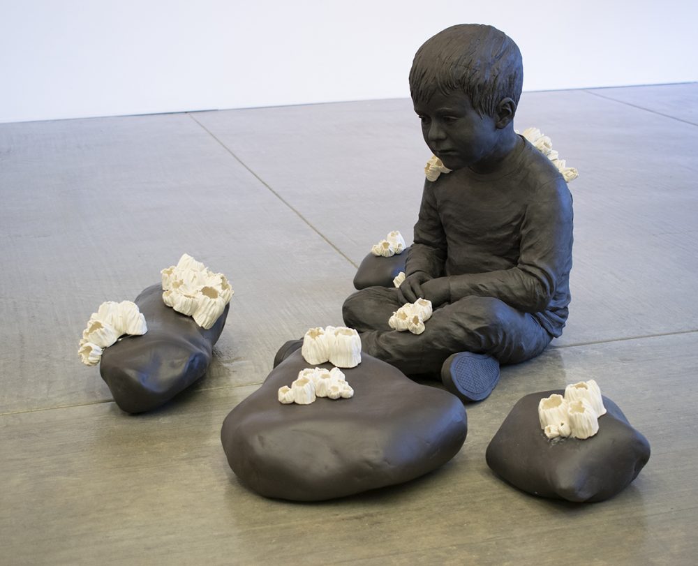 Sculptural installation made of stoneware and porcelain, placed on a concrete gallery floor, with a white gallery wall in the background. The installation features a sculpture of a lifelike depiction of a small boy sitting crosslegged on the floor. There are four large stones arranged around him. The boy and stones are monochromed in a dark brown color. On top of the stones and the boy are sculptures of white barnacles, also realistically rendered. The boy is looking at the stones.