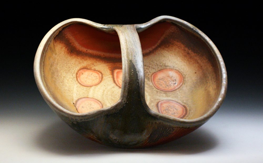 The image is of a ceramic bowl that is shaped like a low, wide basket. There is a handle at the center of the basket shape, and the inside is decorated with orange circle shapes. The glaze on the ceramic is a light brown. The artwork sits at an angle to reveal the decorative interior. The background is white at the bottom of the photo and fades slowly to black as it goes to the top.