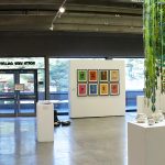 Panorama view of gallery with several artworks installed. At the left are a series of small prints in black frames hung on a white wall. Further along the wall as it recedes into the background are other framed artworks and a small red and blue sculptural element along the wall on the floor. At the far right of the image is a pedestal with a ceramic sculpture on top, and a small wall with framed pieces. The back wall has several multi-colored paintings and other wall two dimensional pieces. Several pedestals are placed throughout the gallery. Two smaller walls are in the center background, one with a colorful series of eight prints, the other with a single wall mounted artwork. Hanging from the ceiling in the center of the room is a sculptural installation consisting of many strips of paper with green leaf shapes.