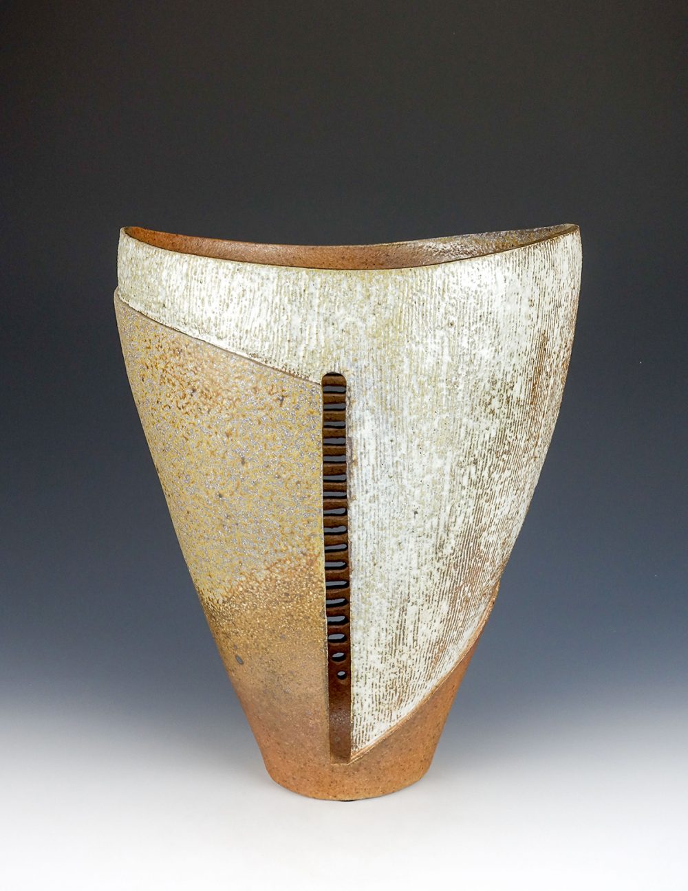 A ceramic vessel sits on a white surface, in front of a grey background. The vessel is a V-shape, with a narrow rounded bottom, and sides that rise steeply as they go up. The opening at the top arcs down in the middle. At the center of the piece is a vertical opening that reveals a series of horizontal openings in the wall of the form. The design features a striated white area on the right that moves across to the left at the top of the piece. The rest of the left side is a mottled beige and ochre color and the bottom is a rusty orange.