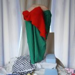 Image Description: The frame is filled with varying fabrics. The background is curtained with white and creme, the floor is white with blue, yellow, and checkered pieces. An obscured body stands in the center. A pillar of green with a center of bright red, a white body is hunched and skin protrudes from the top, the body part is unknown.