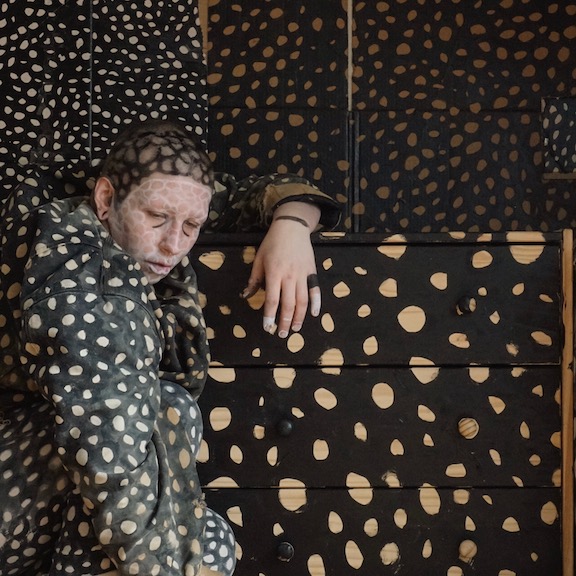 Image Description : A background made of cardboard is painted with interlacing black circles creating a net-like pattern. A dresser made of light wood sits in front painted with the same pattern. A white agender person sits hunched over the dresser on the lefthand side of the image. They are wearing a shapeless canvas coat painted with the same pattern. Their face is painted with white interconnected circles with black lines under their eyes. They have a shaved head which is painted with the black circular pattern. Their hand rests atop the dresser where their wrist and fingers and painted with black and white bands.