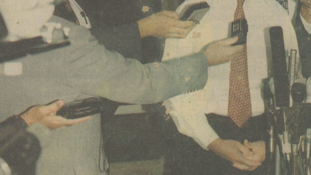 A video still of Untitled (Saturday, October 16, 1993) shows a weathered newspaper clipping of a figure dressed in a white shirt with red tie standing before microphones. Arms reach out from the left side of the shot with tape recorders and additional microphones preparing to record the speaker’s voice.