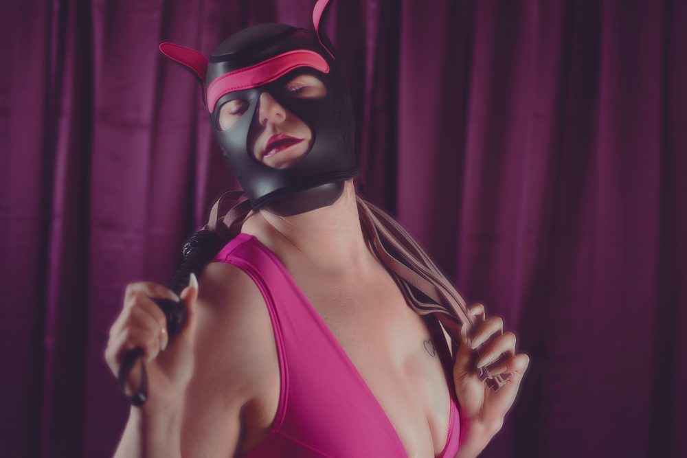 Woman in BDSM cat mask with pink body suit and whip stretched over her back.