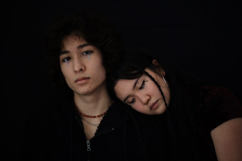 Two people standing in front of a black background. The man on the left is looking at the camera with blue gemstones coming down his eyes. There is a woman resting her head on the man's shoulders. There is glitter, tears, and gemstones coming down her eyes.