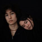 Two people standing in front of a black background. The man on the left is looking at the camera with blue gemstones coming down his eyes. There is a woman resting her head on the man's shoulders. There is glitter, tears, and gemstones coming down her eyes.