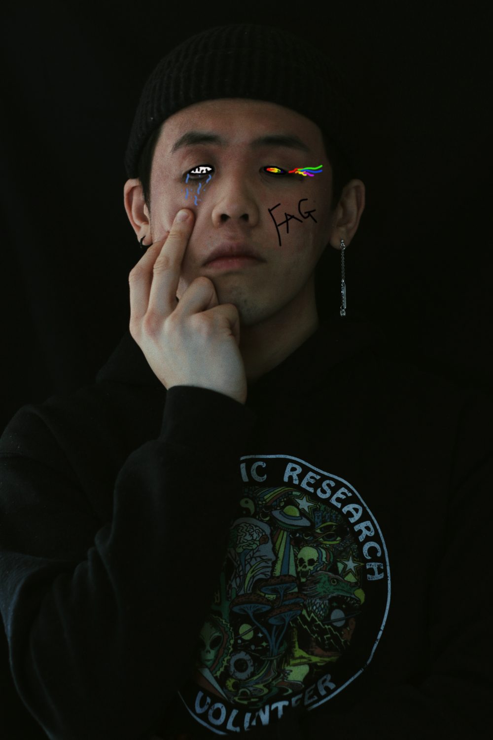 A young man standing in front of a solid black background. Rainbow tears are graphically drawn coming out of the right eye, and there are blue tears coming out on the left. Model is posed in a neutral stance with middle finger pulling down the eye with blue tears. "F*g" is written on his face.